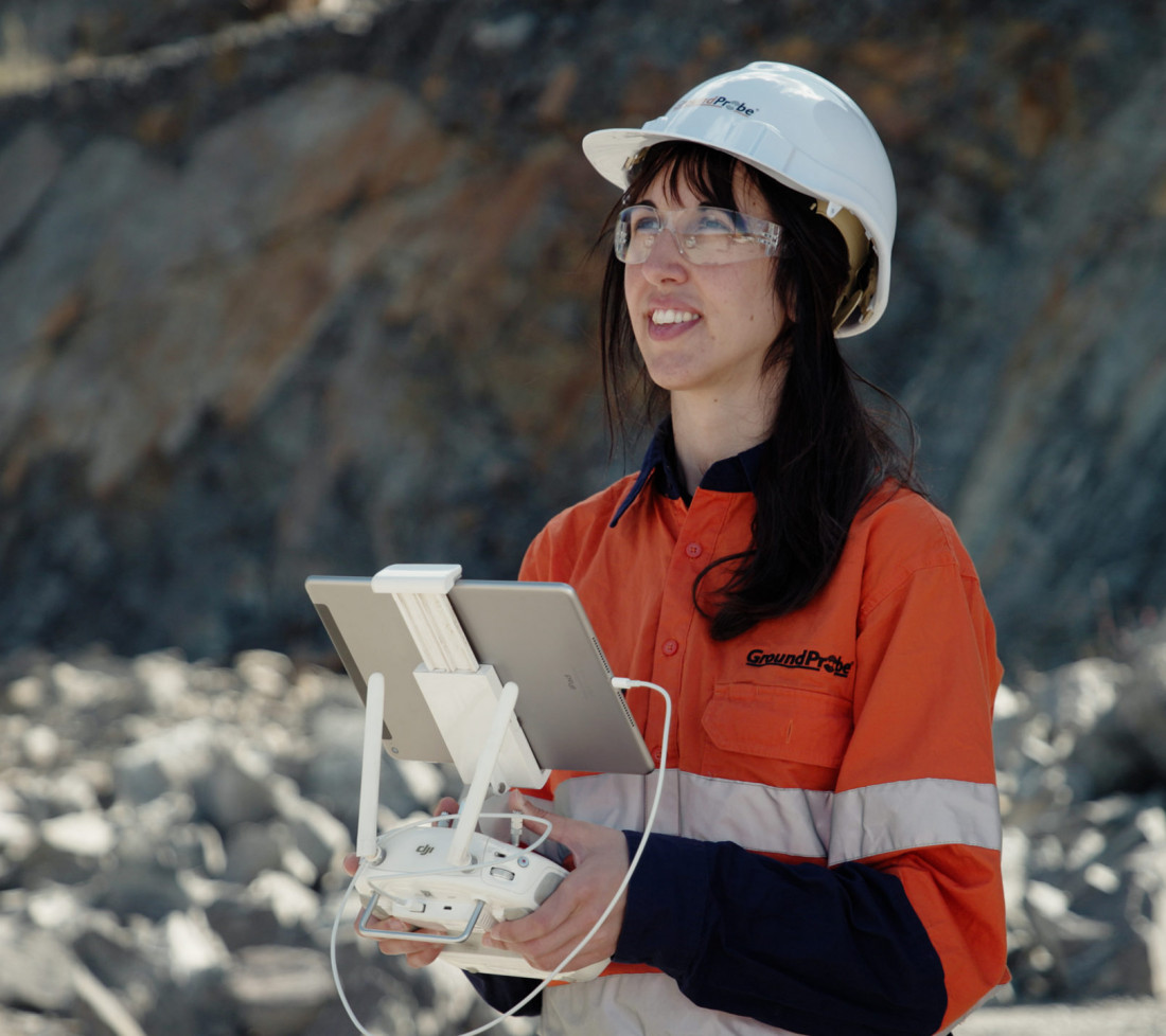 A close-up of a female GroundProbe employee in HPPE gear flying a drone using a tablet and remote controls