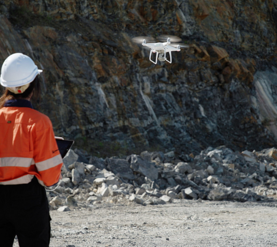 A GroundProbe employee wearing safety gear and flying a drone above a mine site using a control tablet