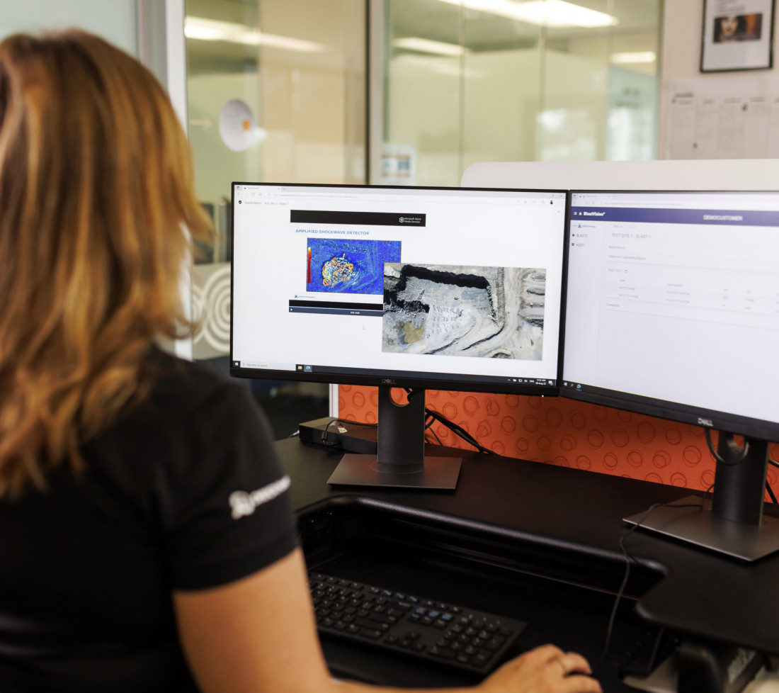 A side view close-up of a GroundProbe female employee sitting at a desk with multiple computer monitors using the GroundProbe BlastVision software to monitor a mining site