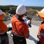 Three miners with their backs to the camera are wearing neon orange construction gear and pointing out towards a mining site.