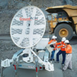 Three GroundProbe geotechnical experts are talking together in orange safety gear while standing beside a large white Groundprobe circular radar. 
