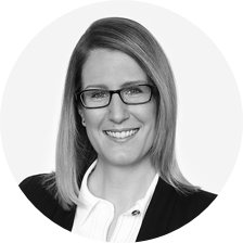 a black and white photo of a woman with long blonde hair and black glasses posing for a company head shot.