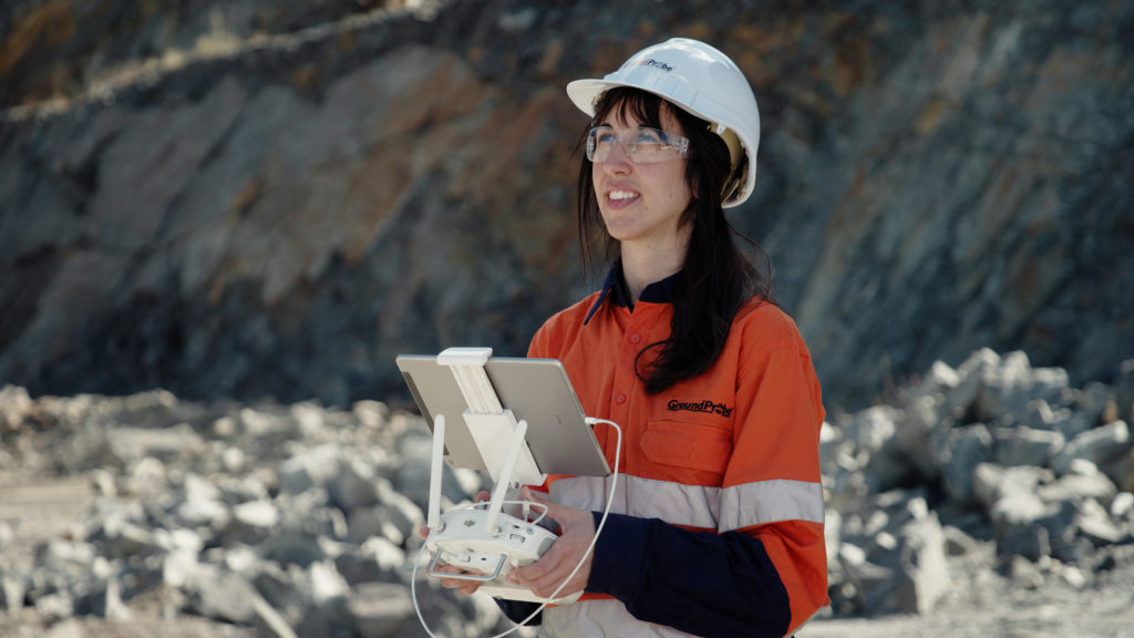 A close-up of a female GroundProbe employee in HPPE gear flying a drone using a tablet and remote controls