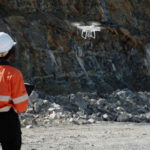 A GroundProbe employee wearing safety gear and flying a drone above a mine site using a control tablet